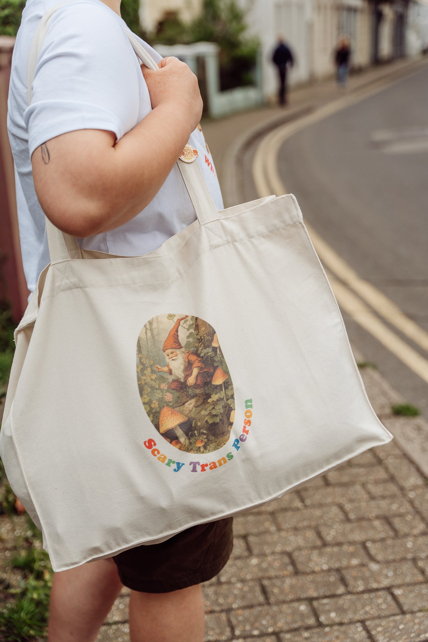 Person standing on a street and holding a beige tote bag with a woodland gnome graphic and 'Scary Trans Person' text