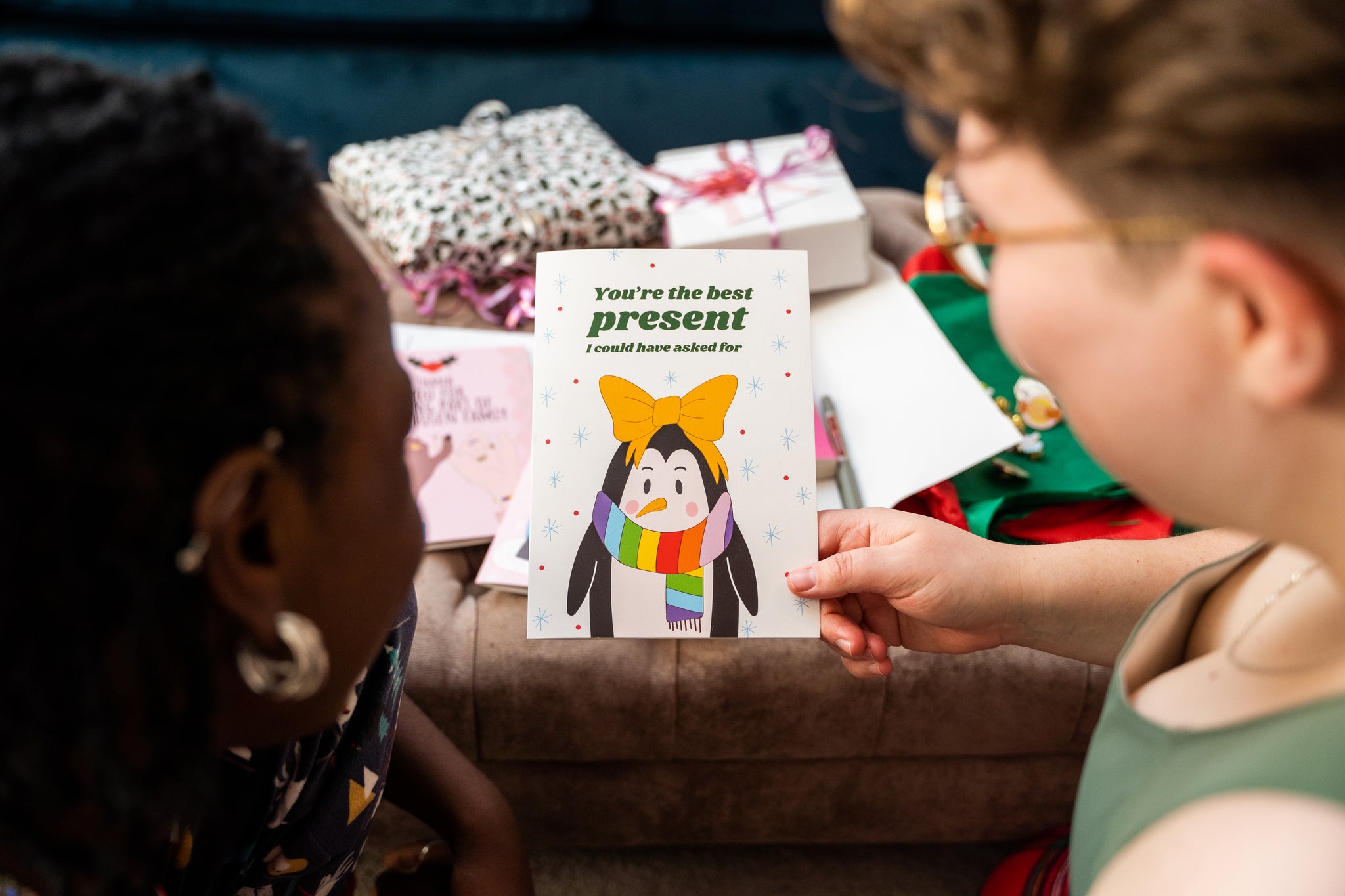 Two people looking at a card reading 'Youre the best present I could have asked for' with a penguin and rainbow design, surrounded by gift boxes on a couch.