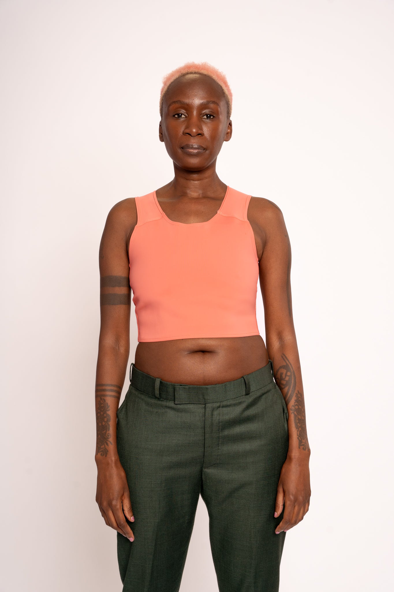 Person wearing a coral coloured short chest binder and dark green trousers  against a white background