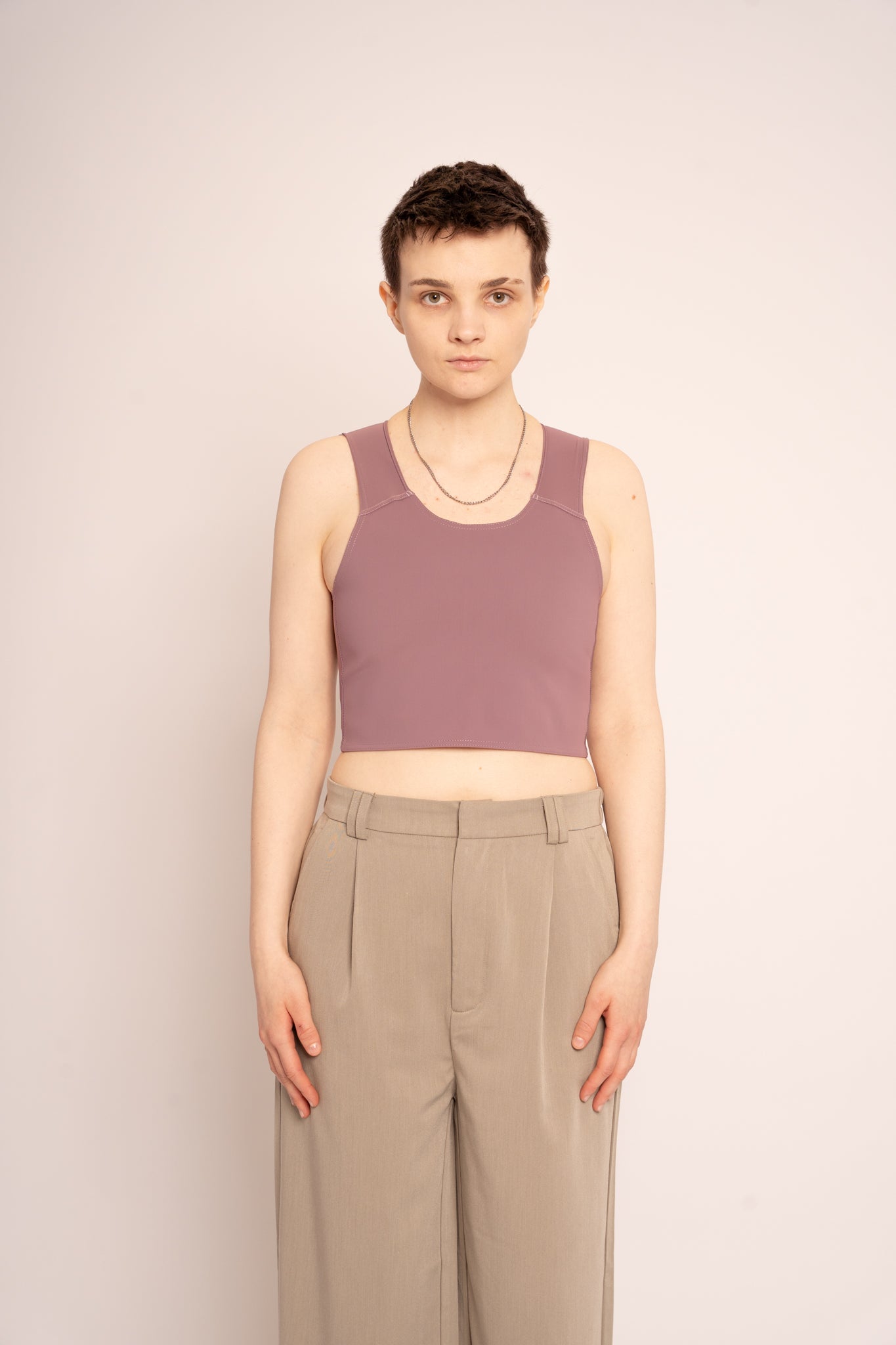 Person wearing a fig coloured short chest binder and beige straight leg trousers against a white background