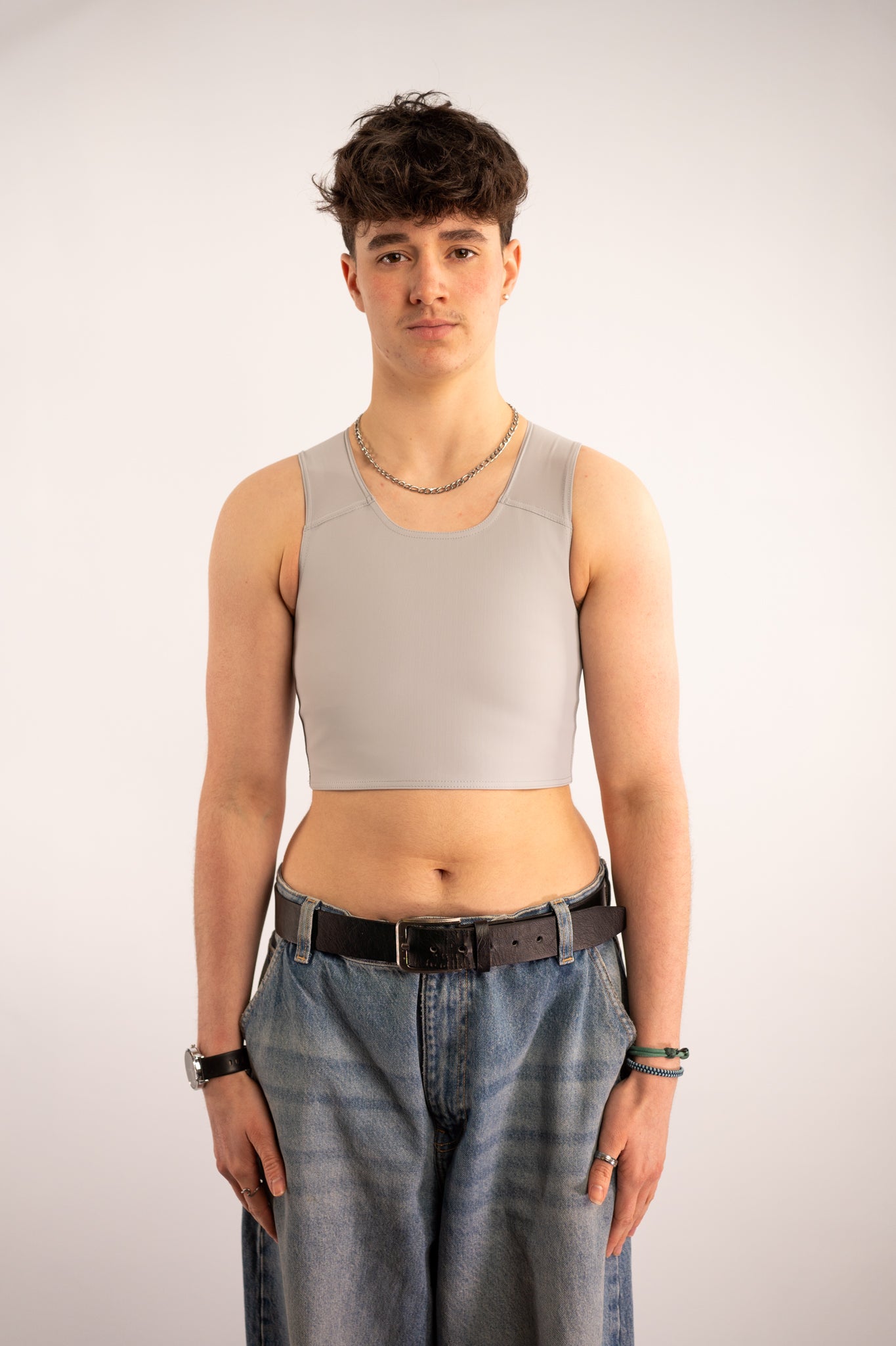 Person wearing a grey coloured short chest binder and blue jeans with a black belt against a white background