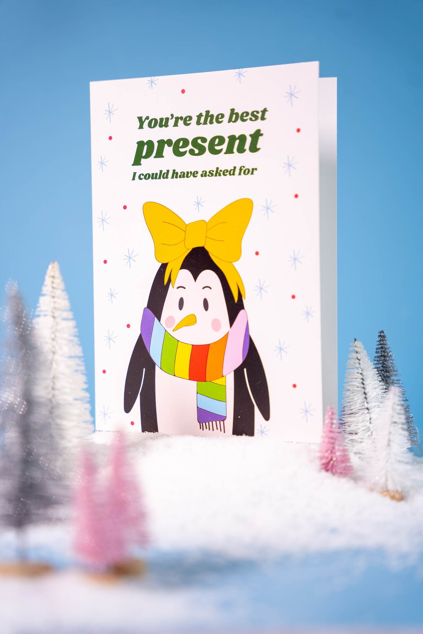 Card with text "You're the best present I could have asked for' and a penguin wearing a rainbow scarf and yellow bow, surrounded by Christmas trees on a blue background.