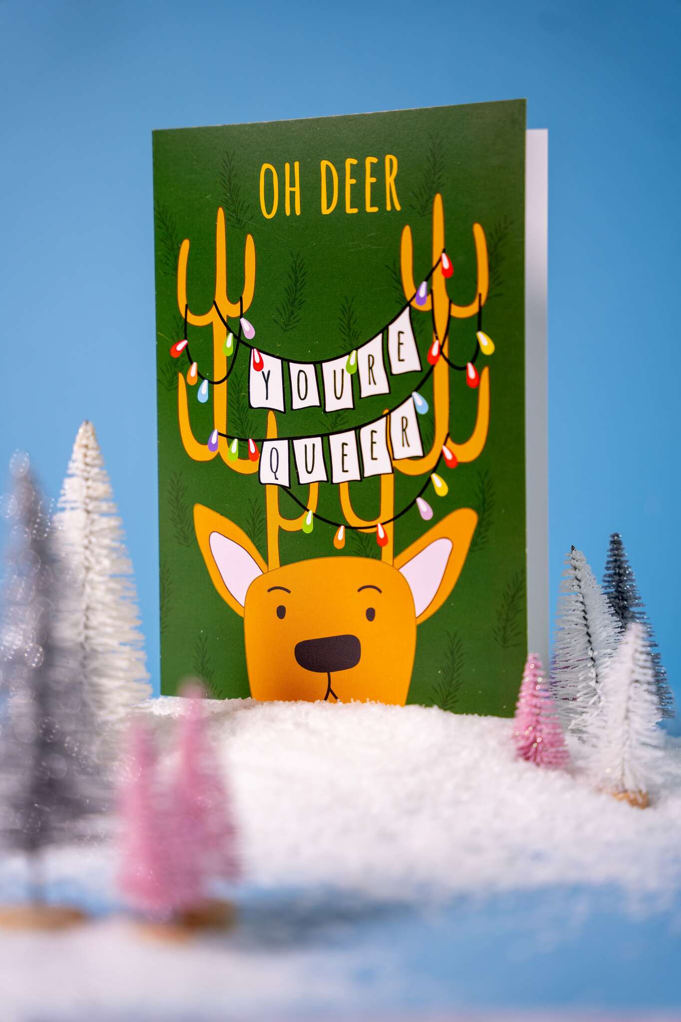 Card with a deer wearing antlers with lights and text 'Oh Deer You're Queer' on a snowy landscape background.