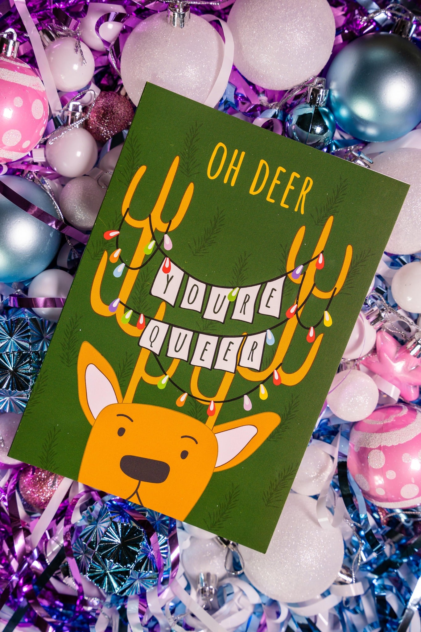 Card with a reindeer illustration and text 'Oh Deer, You're Queer!' on a festive background with colorful ornaments.
