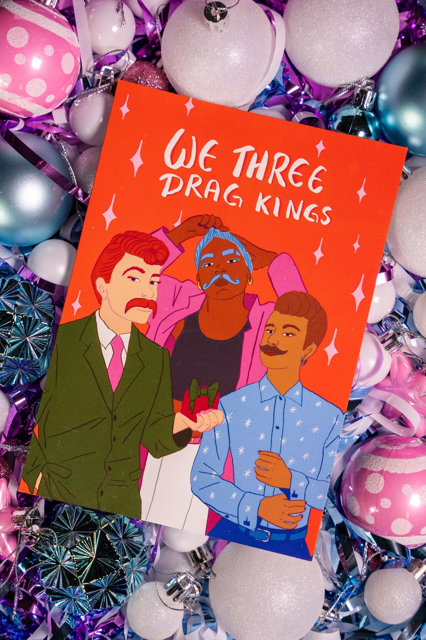 Card with illustration of three drag kings on an orange background, surrounded by festive decorations.