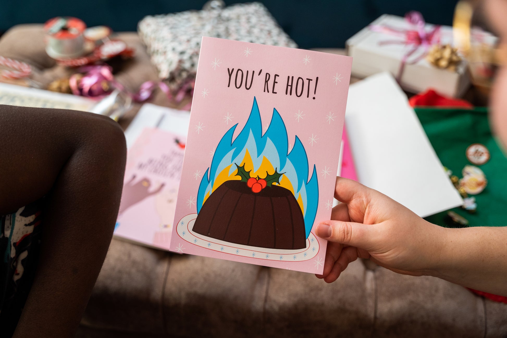 Card with a flaming christmas pudding illustration and 'You're Hot!' text held by a person in a festive setting.